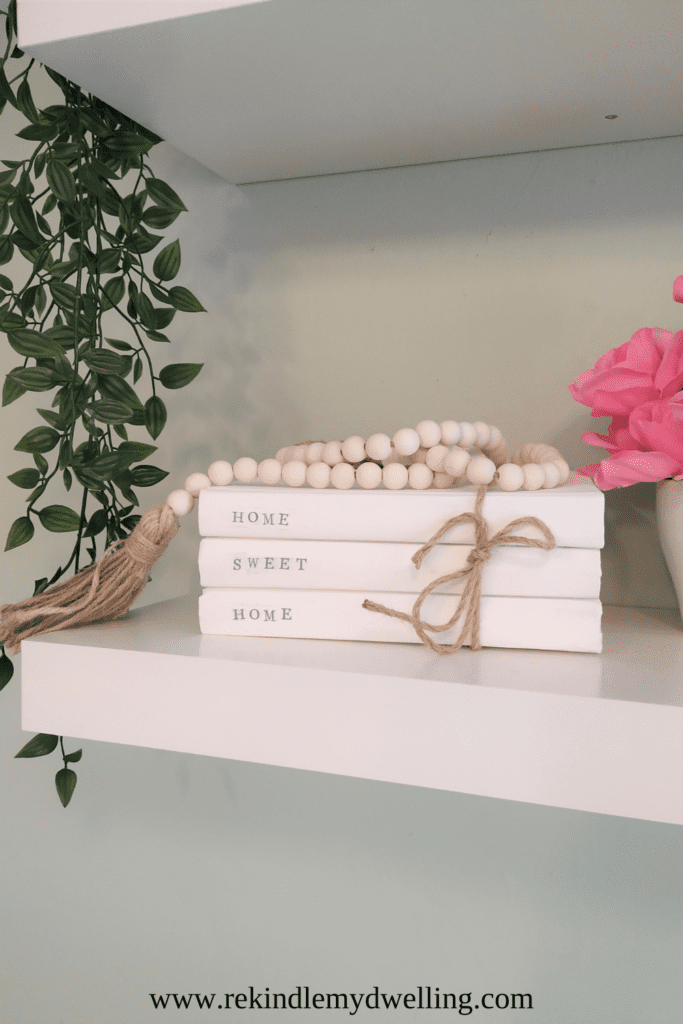 Stacked book decor on a shelf next to beads, greenery and pink flowers.