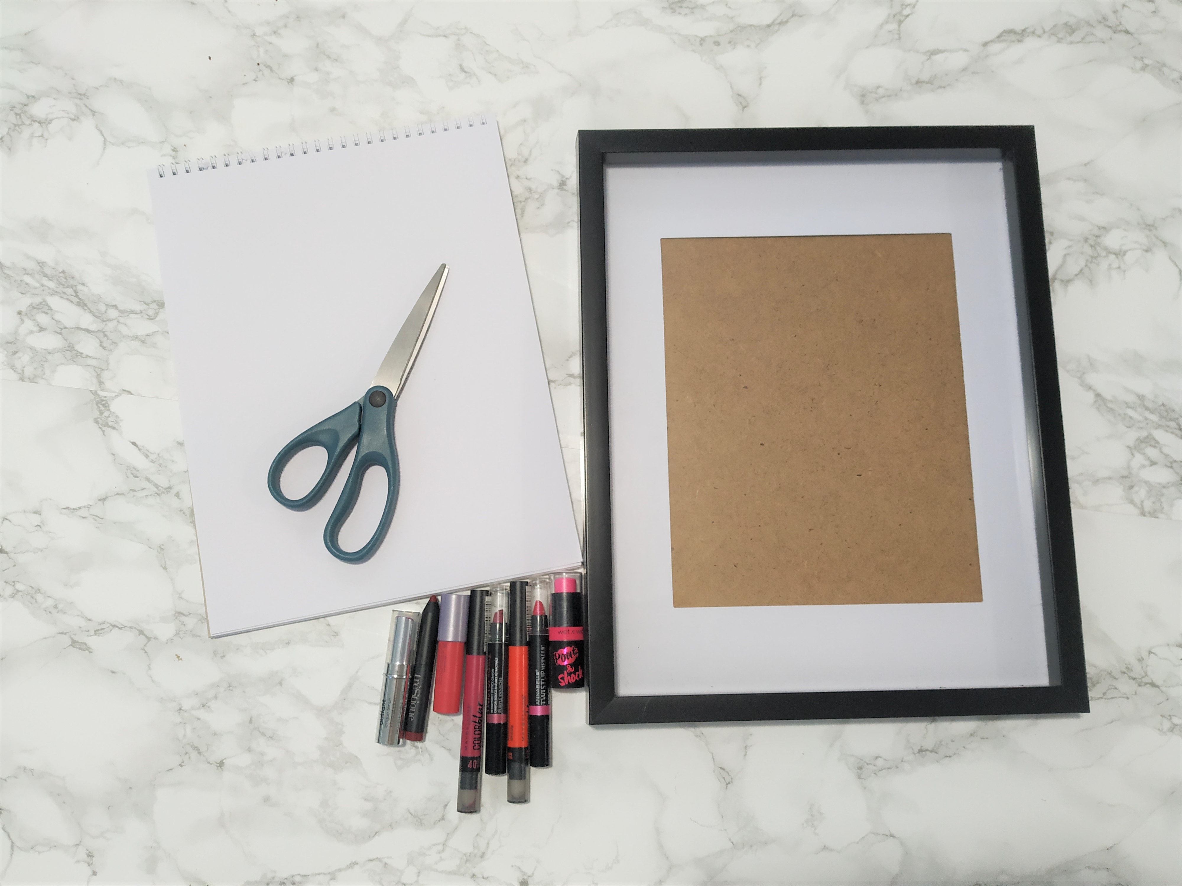 Materials needed to make lipstick art including paper, scissors, picture frame and lipstick.
