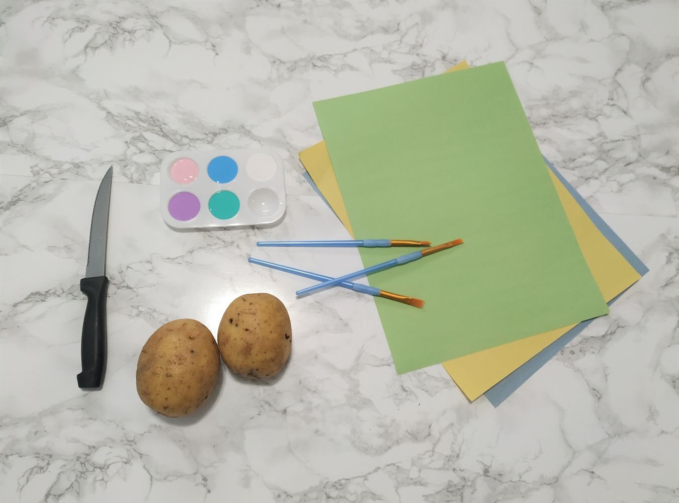 Materials needed to make potato stamp Easter eggs including potatoes, a knife, paint, paintbrushes and paper.