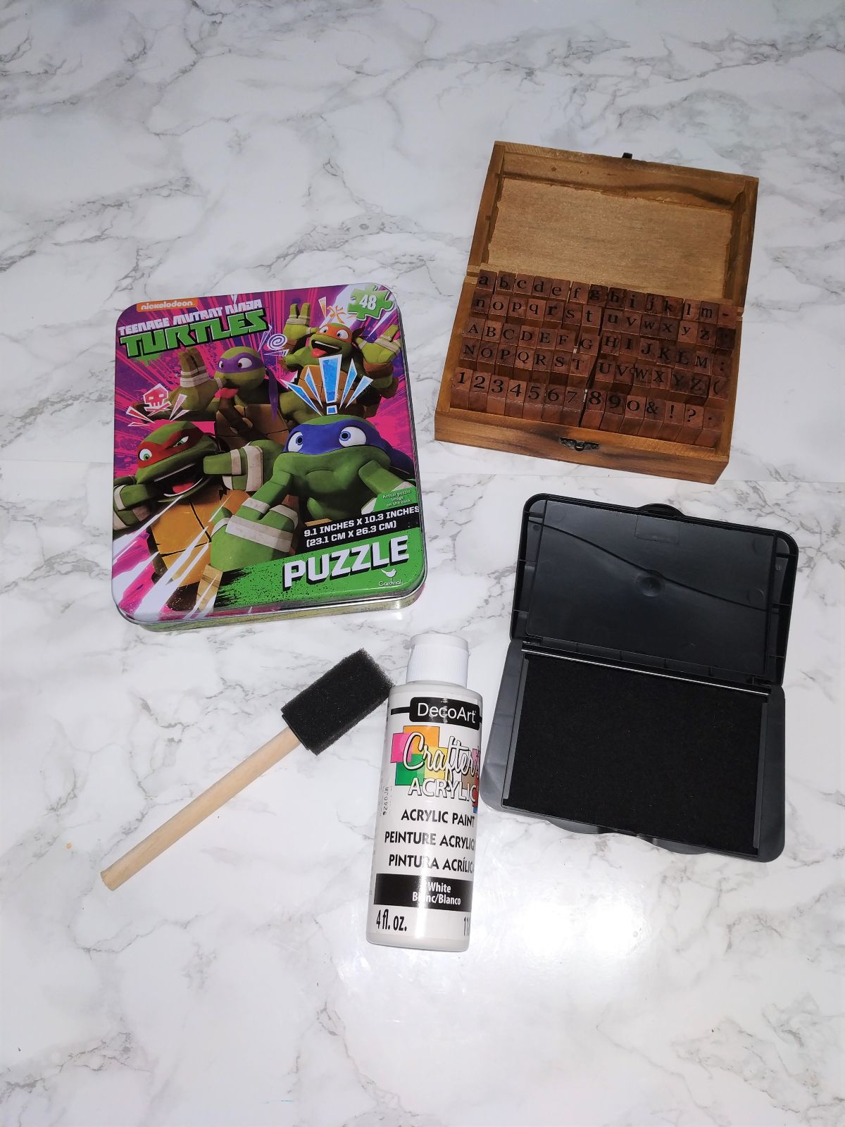 Materials needed to make a gift box including a tin box, paint, paintbrush and stamps.