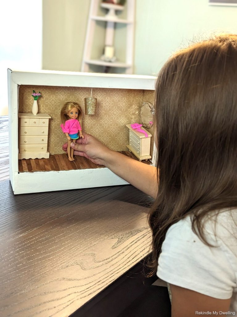 Girl playing with a doll in a diy cardboard box dollhouse.
